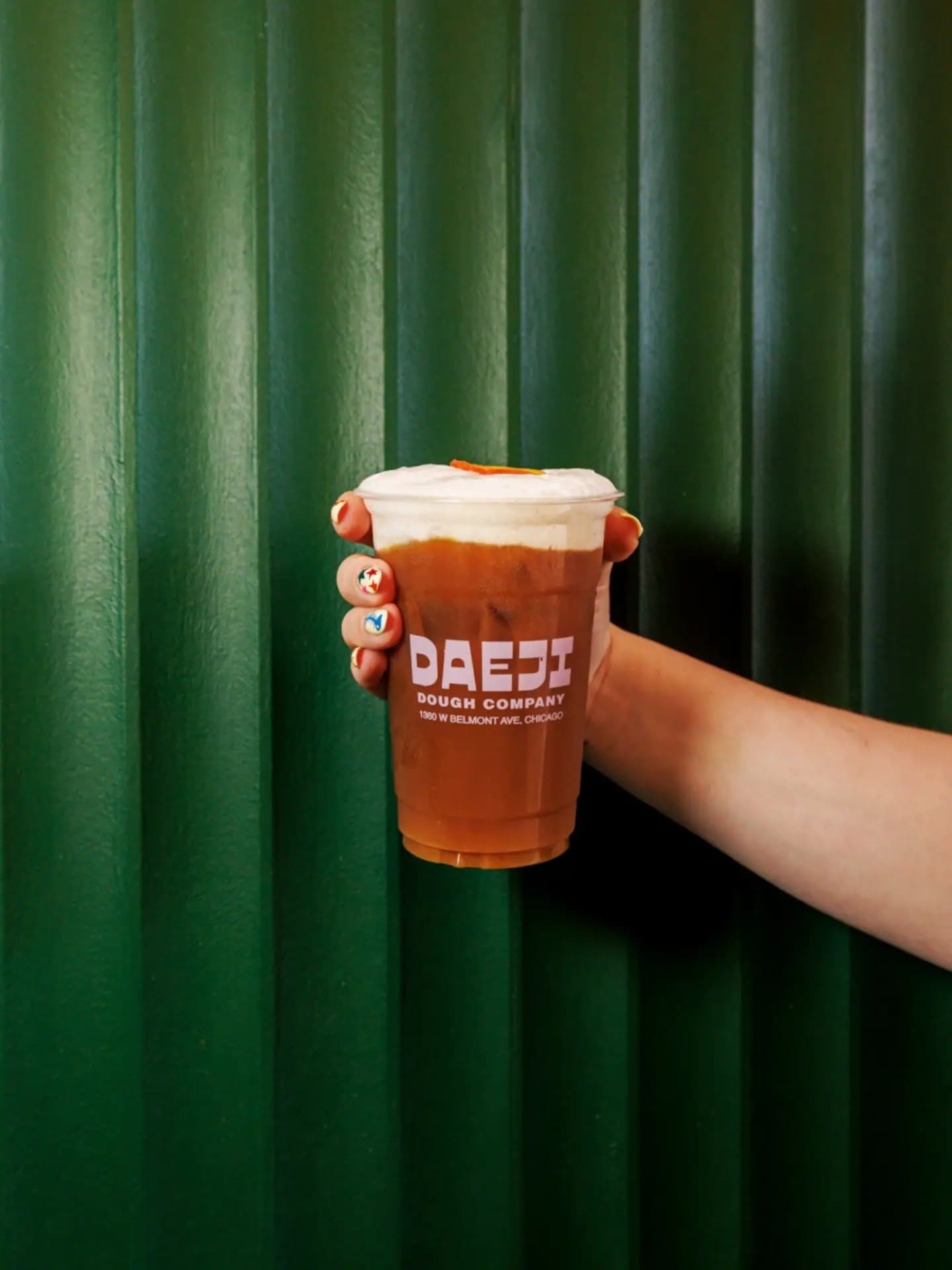 A hand holding a Daeji Dough Co cup in front of a green curtain